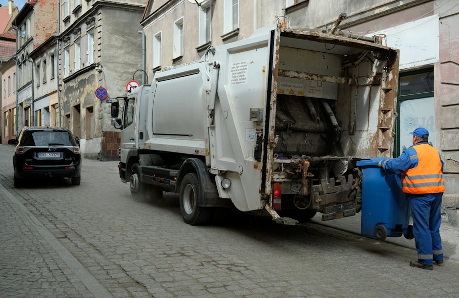 A street scene featuring a white waste collection truck with rust and dirt visible on its rear compartment, actively engaged in rubbish removal, with a worker in a high-visibility orange vest and blue uniform positioned beside the truck, handling a blue wheeled bin. The truck is parked adjacent to old, multi-story buildings with weathered facades, including stone and brick finishes. The cobblestone street is narrow, with a black car parked further down the road. The truck's rear is open, revealing mechanical components and an empty compartment prepared for rubbish collection. Environmental lighting appears natural, with daylight illuminating the scene, emphasizing the urban environment and the process of private waste disposal that may be an alternative to council-provided rubbish collection services, aligning with the context of independent rubbish removal efforts by companies like House Clearance Harringay.
