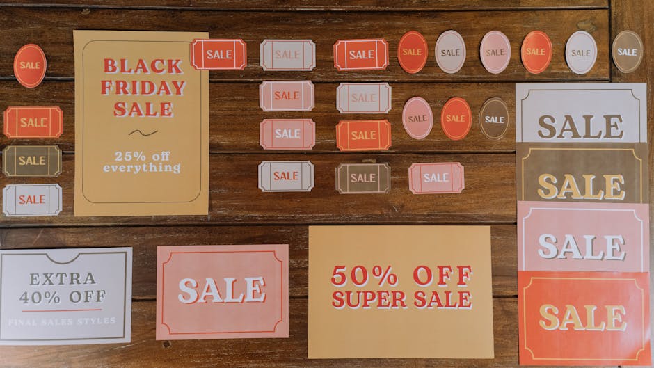 A collection of various sale signs displayed on a wooden surface, including rectangular, oval, and circular shapes in shades of red, white, pink, and beige. Prominent among them are signs with bold text such as 'BLACK FRIDAY SALE' indicating a 25% discount, and another with '50% OFF SUPER SALE' on a beige background. Smaller tags labeled 'SALE' are arranged in rows, some with decorative borders and others plain, with one sign offering an 'EXTRA 40% OFF FINAL SALES STYLES' in a white and grey design. The signs feature different textures, with some resembling paper materials with matte and semi-gloss finishes. The wooden surface has a warm, medium-brown tone with visible wood grain, providing a natural background. The lighting appears soft and even, illuminating the signs without harsh shadows, emphasizing the promotional content for sale events commonly used in retail displays for clearance or promotional clearance sales, reflecting on the themes of discount advertising and retail marketing. The arrangement includes signs with varied font styles and sizes, illustrating a typical collection of promotional signage used in shop window displays or clearance sections to attract customer attention to offers and discounts.