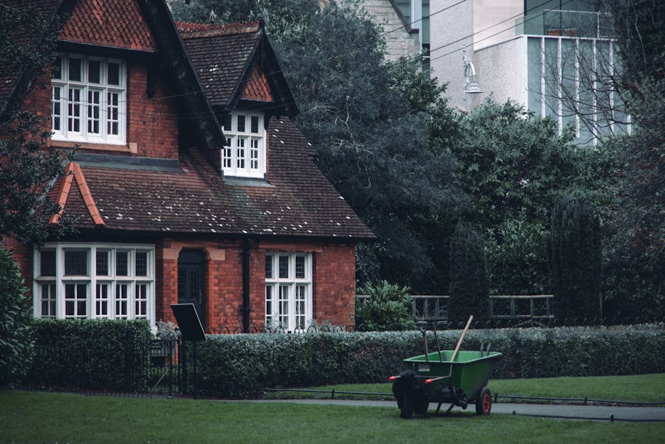 In the foreground of the image, a green wheelbarrow with red handles and black wheels is positioned on a paved pathway, filled with a few wooden poles and partially vacant. The pathway runs parallel to a well-maintained lawn bordered by a low, neatly trimmed hedge. Behind the hedge stands a traditional red brick house with a steep, dark tiled roof sporting multiple gabled sections and a small front porch. The house features white-framed, multi-pane sash windows and a dark-colored front door. To the left and slightly in front of the house, there is a black metal fence with a gate, adjacent to a dark outdoor chair. The upper part of the house shows two small dormer windows within the roof structure. In the background, there is dense greenery, including tall shrubs and trees, and beyond that, sections of modern high-rise buildings with glass facades are visible, indicating proximity to an urban area. The atmosphere appears overcast or early evening, with diffused lighting illuminating the scene. The visual context suggests a residential setting that might involve private waste or garden clearance, consistent with services offered by House Clearance Harringay.
