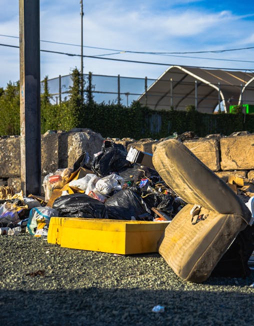 A pile of mixed waste materials, including black plastic trash bags, a yellow plastic container, and a worn, beige car seat cushion, is situated on a gravel surface in an outdoor location. The waste is positioned against a low stone wall composed of large, uneven, grey stone blocks. In the background, there are green bushes, a tall metal pole supporting overhead power lines, and a sports or tennis court enclosure with a high black mesh fence. A large, semi-circular metal-framed structure with a white cover is also visible, along with a clear blue sky with some wispy clouds. The scene suggests an area awaiting rubbish collection or disposal, indicating the need for professional waste removal services by companies such as House Clearance Harringay that handle private rubbish clearance and on-site or alternative waste handling solutions. The overall environment and objects in the image emphasize the importance of proper rubbish removal in maintaining cleanliness and safely managing waste materials.