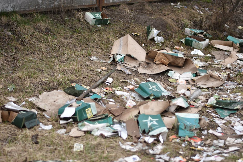 An outdoor scene showing a patch of ground with scattered debris, including disorganized cardboard boxes, some partially collapsed and torn, with visible printed logos and labels, alongside crumpled paper, plastic packaging, and bits of torn cardboard. The ground is covered with patchy grass and dirt, with a slight incline towards the background. In the upper left corner, part of a wooden fence or barrier is visible. The debris appears informal and unmanaged, indicating dumped waste, possibly from packaging materials. The lighting is natural, suggesting daytime, and the overall scene reflects improper disposal of waste, aligning with services offered by companies like House Clearance Harringay who undertake alternative waste handling or private rubbish collection.