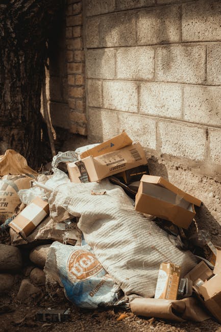 A collection of discarded cardboard boxes and paper waste piled against a weathered brick wall, with some boxes partially crushed or torn. The boxes vary in size and are mostly brown, with a few featuring printed labels or barcode markings. Among the rubbish, there are also black plastic bags and large white sacks filled with unknown materials, placed on rough, uneven ground with scattered small stones and debris. The scene is outdoors, adjacent to a brick and concrete wall that appears aged, with visible mortar joints and weathering. A large tree trunk is visible on the left side of the image, casting shadows over part of the rubbish. The lighting is natural, suggesting daytime, emphasizing the textures of the cardboard, bags, and the wall surface. This scene exemplifies typical private waste accumulation that could be addressed through independent rubbish removal services, such as those offered by House Clearance Harringay, rather than local authority collection.