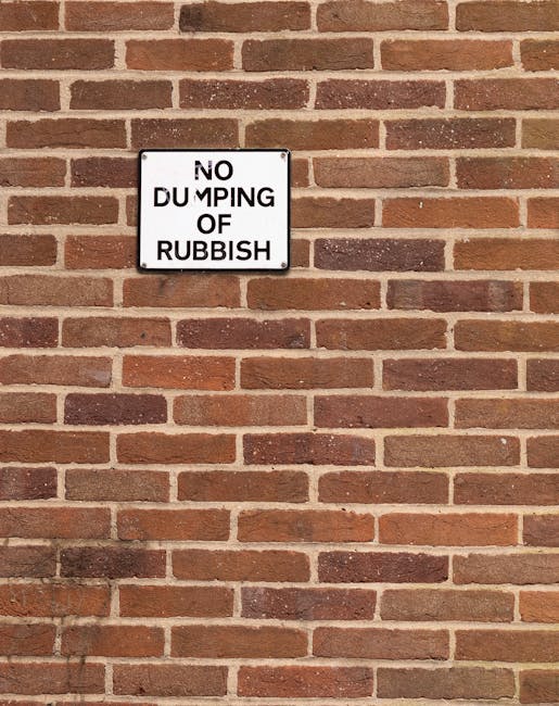 A rectangular white and black metal sign mounted on a red brick wall displaying the message 'NO DUMPING OF RUBBISH'. The bricks are arranged in a horizontal pattern with visible mortar joints, and the wall has a slightly weathered appearance with some variations in colour and texture. The sign is centered in the upper left section of the image, with the brick surface occupying the rest of the view. The lighting is natural, casting soft shadows on the brick surface, highlighting the rough texture of the bricks and the smooth finish of the sign, which serves as a visual reminder of waste disposal regulations in the area. The scene relates to waste management and illegal rubbish dumping prevention, consistent with a professional waste removal service such as House Clearance Harringay, emphasizing the importance of proper disposal to avoid fines or penalties.