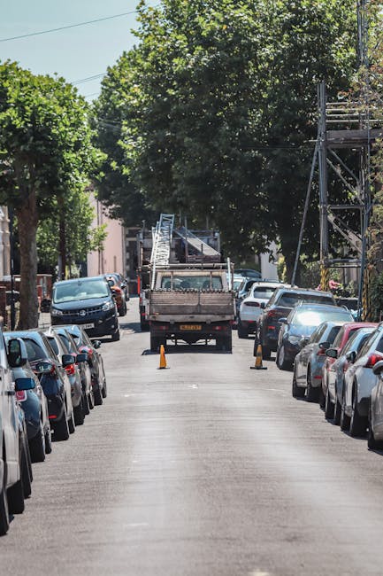 A narrow urban street lined with parked cars on both sides, including compact and sedans with metallic and dark-coloured finishes, is the primary scene in the image. In the middle of the road, a flatbed truck with a metal frame and wooden sides is positioned, surrounded by orange safety cones indicating ongoing debris removal or clearance work. The truck appears to be engaged in a waste collection process, possibly involving rubbish or building materials, consistent with private waste handling or site clearance activities. The street is shaded by mature trees with full green foliage, casting dappled sunlight across the scene and enhancing the environment's natural urban character. In the background, residential buildings with brick facades are partially visible, while overhead, power lines and a streetlight support the cityscape context. The overall lighting suggests a clear day, and the scene subtly reflects an alternative method of rubbish removal outside traditional municipal collection, indicative of independent waste disposal or on-site clearance services provided by companies such as House Clearance Harringay.