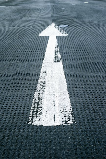 A close-up view of a textured dark grey surface, likely a parking or loading area, featuring a large white arrow painted centrally pointing straight ahead. The arrow's broad head and straight shaft are clearly visible, with slightly worn edges and some minor chipping of the white paint. The surface underneath has a grid-like pattern with small raised bumps, providing traction. The background extends into a slightly lighter area with a hint of natural light, suggesting outdoor surroundings. This scene could relate to waste management and independent collection routes, indicating directional guidance for vehicles or service personnel. House Clearance Harringay provides professional rubbish removal services, often operating in such paved or industrial environments, where clear marking assists efficient on-site clearance and waste collection activities. The image exemplifies how marked pathways facilitate orderly rubbish handling and disposal processes, supporting effective private waste management practices in urban or residential areas.