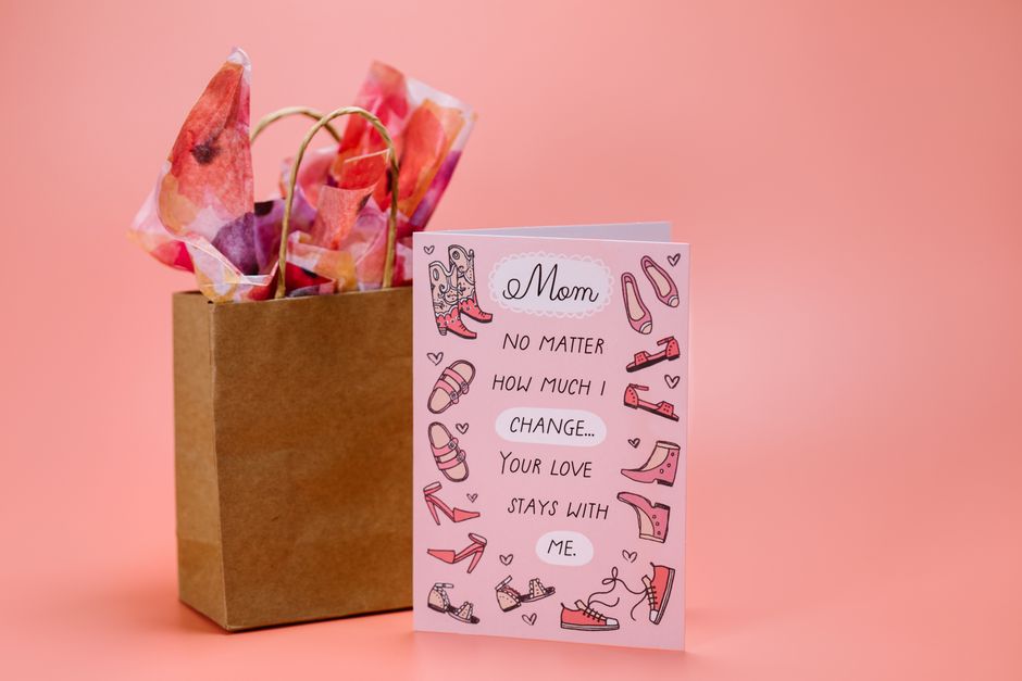 A brown paper gift bag with twisted paper handles sits upright on a flat surface, containing various tissue or wrapping paper in shades of pink, peach, and purple, with some slightly crumpled or folded. Positioned in front of the bag is a greeting card with a pink background, decorated with illustrated pink and purple shoes, including heels, trainers, and sandals, scattered around the message. The card's central text reads, 'Mom no matter how much I change... your love stays with me,' written in black and pink ink, with small heart illustrations interspersed among the shoes and text. The scene is set against a soft, solid pink backdrop, emphasizing the gift and card with gentle lighting that creates subtle shadows, suggesting a studio setting focused on the theme of Mother's Day or a special occasion involving thoughtful gift-giving related to family and feminine accessories. This arrangement supports themes of personal connection and thoughtful preparation, relevant to private gift or waste management services for personal occasions.