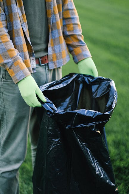 A person wearing a yellow and grey checked shirt, green gloves, and grey trousers is standing outdoors on a grassy area, holding open a large black plastic rubbish bag. The bag is empty and held open at the top, ready for waste disposal. The background shows a well-maintained lawn, suggesting the scene is taking place in a garden or park setting. The person's torso and arms are visible, with the focus on the act of preparing the rubbish bag for waste collection, which relates to the context of waste management and rubbish removal services. The scene emphasizes proper handling of rubbish in an outdoor environment, consistent with private waste disposal or rubbish clearance activities conducted by services such as House Clearance Harringay, supporting the process of independent collection or on-site clearance in a professional manner.