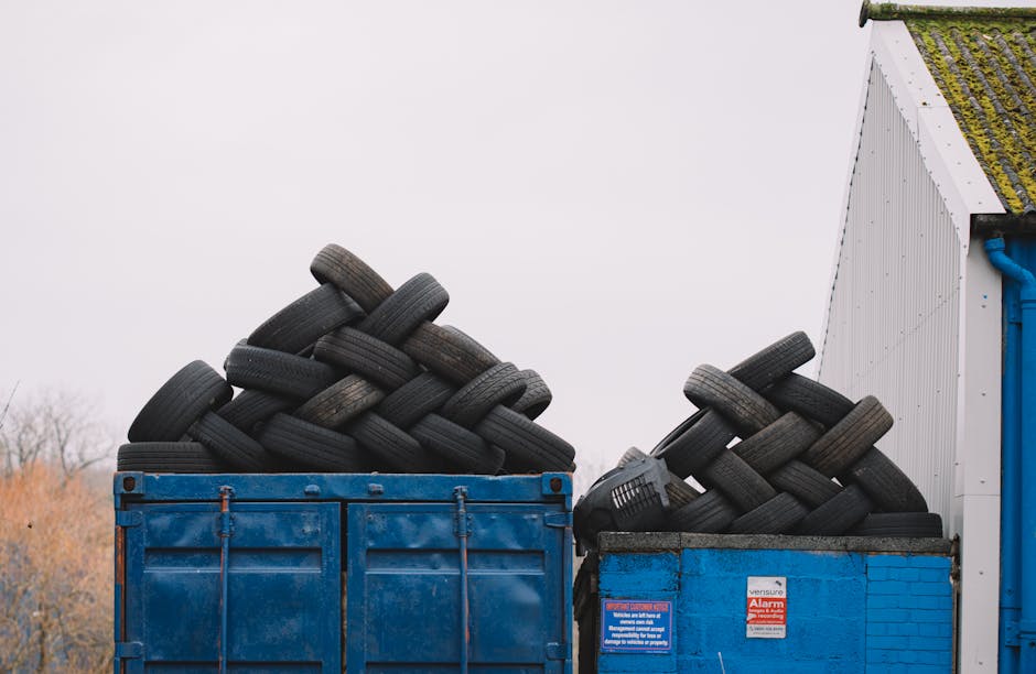 A large blue industrial skip positioned outdoors on a concrete surface, filled with a significant pile of used black rubber tires stacked haphazardly with some leaning against the edges of the container. The tires vary slightly in size but are predominantly similar in appearance, exhibiting textured rubber surfaces and visible tread patterns. Adjacent to the blue skip, on the right, is a smaller blue brick container also filled with tires, with an orange alarm sign attached to its side. In the background, a section of a white building with green moss accumulation on the roof and a corner of a blue pipe is visible, while the sky overhead is overcast, casting diffuse natural light across the scene. The setting appears to be an outdoor yard space used for waste or recycling management, aligning with private waste handling practices associated with rubbish removal services.