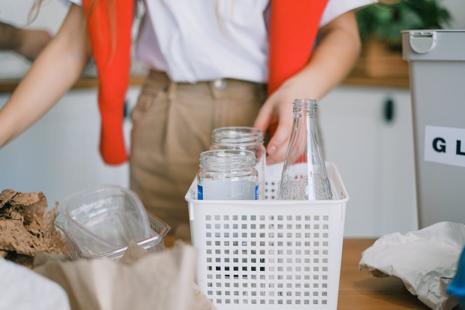 A person wearing a white shirt and beige trousers is standing at a workspace, placing several empty glass jars into a small white plastic basket with a grid-like pattern on the side. The jars are clear, with some having metal screw lids, and appear to be clean and suitable for recycling or donation. To the left, there is a pile of crumpled brown paper, and a transparent plastic container holding more materials. The background features a light-colored, possibly laminated surface and a wall with cabinets or storage units, with a portion of a gray recycling bin visible on the right side, labeled with text partially out of frame. The lighting is natural and bright, suggesting a well-lit indoor environment focused on sorting or preparing waste for disposal or reuse, consistent with waste management or recycling activities, as often handled by companies such as House Clearance Harringay.