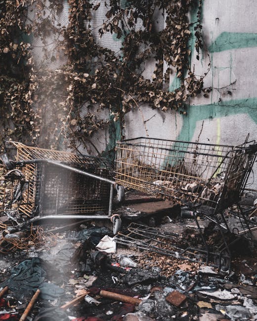 A collection of abandoned shopping trolleys stacked and scattered on a grime-covered concrete surface outdoors, with one trolley overturned and others leaning against a weathered, graffiti-marked wall. The trolleys are made of metal wire, showing rust and corrosion, and are surrounded by burnt debris, charred paper, plastic waste, and ash, indicating recent or ongoing waste disposal or clearing activities. Behind the trolleys, the wall features peeling paint and faded green and grey graffiti, with dried, shriveled vine-like plant growth climbing over it, adding a neglected urban environment feel. The overall scene is shaded and slightly smoky, emphasizing an area where private rubbish or waste could be temporarily accumulated for collection. The setting aligns with the typical context of independent rubbish removal or on-site waste clearance, as provided by services like House Clearance Harringay, emphasizing the importance of professional rubbish removal to address such clutter in communal or commercial outdoor spaces.